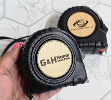 Load image into Gallery viewer, Black 25 ft measure tapes, with wood block circle,engraved with company logos.
